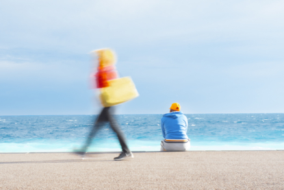 A woman passes by leaving only a blur while a man with an orange cap watches the waves on the Promenade des Anglais in Nice. Days with strong winds make the light and colours very intense. The spectacular blue of the sea stands in contrast with the colours of the people's clothes on a windy afternoon.