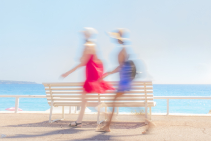 Two ladies dressed in their colourful summer dresses walk past one of the typical white benches on the Promenade des Anglais in Nice, ready to enjoy a day at the beach (September 2017).